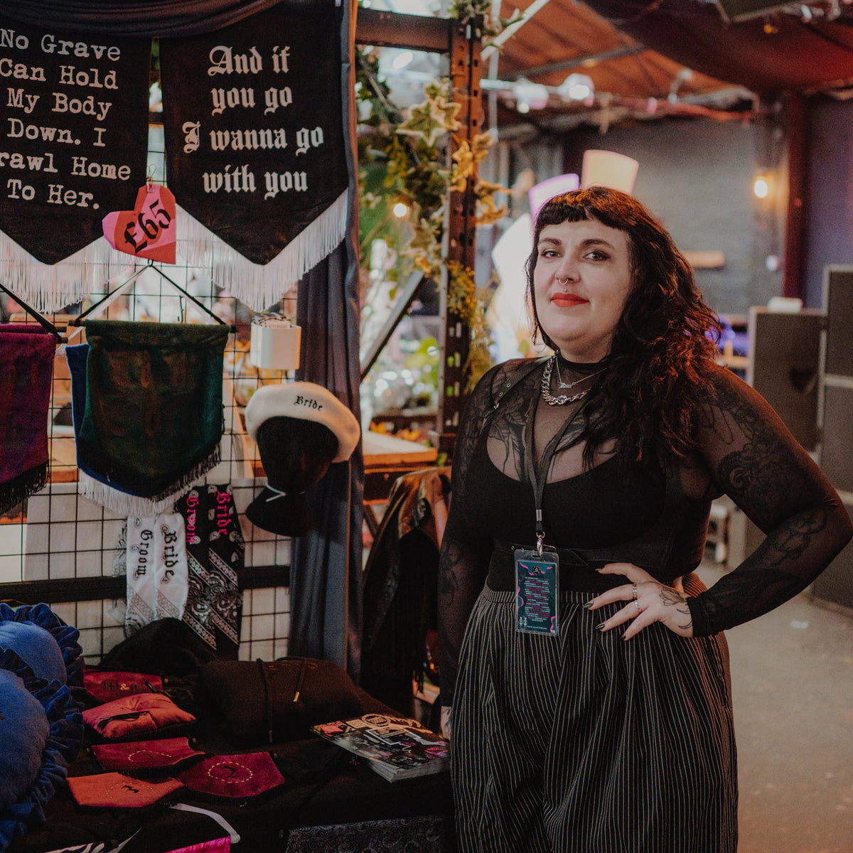 A woman stands at a craft fair booth wearing a sheer black long-sleeve top and dark pinstriped trousers. The booth displays handmade items including a black banner with white gothic text, a black mesh bag, and other apparel under warm overhead lighting.